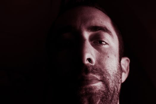 Dramatic portrait of a young man half expressionless face in deep shadow looking at camera isolated on black background.