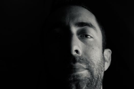 Dramatic portrait of a young man half expressionless face in deep shadow looking at camera isolated on black background.