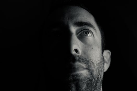 Dramatic portrait of a young man half expressionless face in deep shadow looking up isolated on black background.