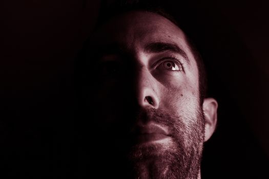 Dramatic portrait of a young man half expressionless face in deep shadow looking up isolated on black background.