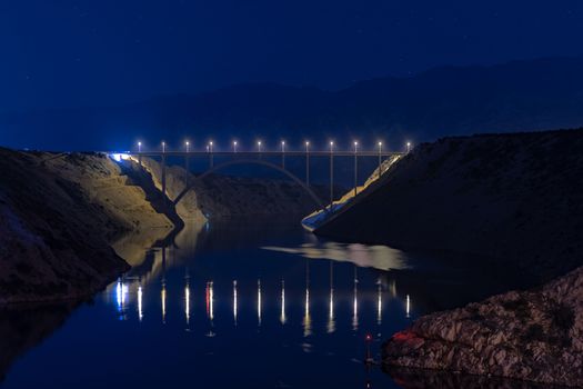 Illuminated highway bridge under stars, reflection of lights in water, mountains in background and bright stars in sky, Maslenica, Croatia