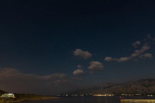 Seascape under stars, ursa major, mountains in background, coast of Croatia at night, pier with lighthouse, Velebit in background, clouds in the sky