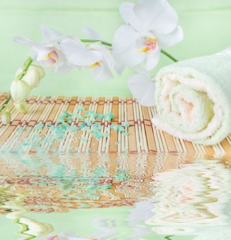 Spa concept: bath towels and bath salt on a natural mat  and a branch of white orchid on a green background reflected in a water surface with small waves