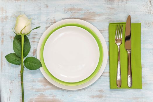 White and green empty plates, green linen napkin, fork and knife and white rose flower on an old wooden table; top view, flat lay, overhead view