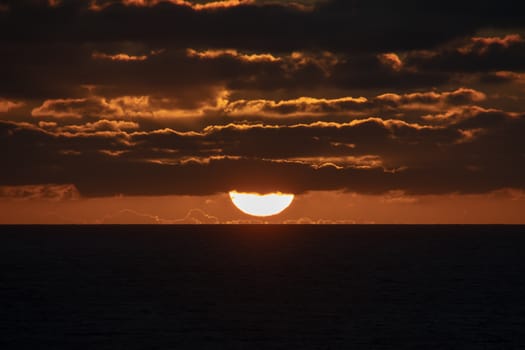 Sun a short moment before touching the horizon partly covered by orange shining clouds