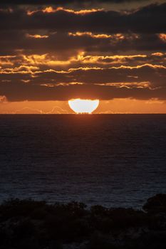 Sun just touching the horizon partly covered by red shining clouds during sunset in Geraldton Western Australia