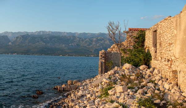 Ruins of stone house on waterfront in sunset, mountains and sea in background, Croatia, town of Vinjerac near Zadar