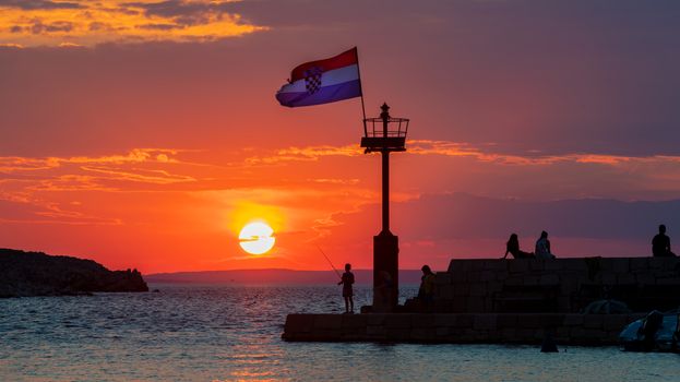 Croatian flag flying in wind at sunset in harbor, colorful clouds over horiyon, boy fishing from pier, girls sunbathing on stone wall, all people silhuetted and unrecognizable