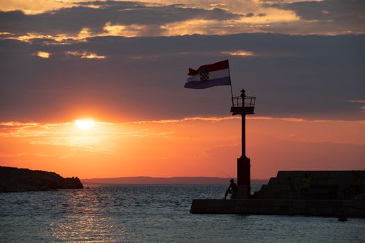 Croatian flag flying in wind at sunset in harbor, man sitting beneath lighthouse watching sunset, all people silhuetted and unrecognizable