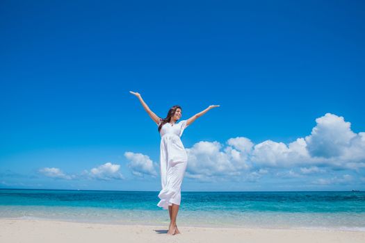 Woman in white dress posing in tropical sea beach with arms raised