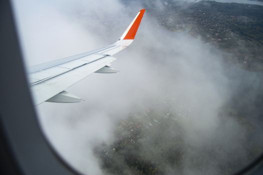 View from the window of the aircraft on the buildings and trees, the wing of the aircraft on the background of passing clouds, the concept of flight.