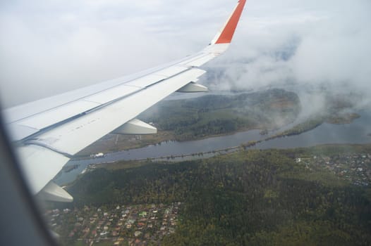 View from the window of the aircraft on the buildings and trees, the wing of the aircraft on the background of passing clouds, the concept of flight.