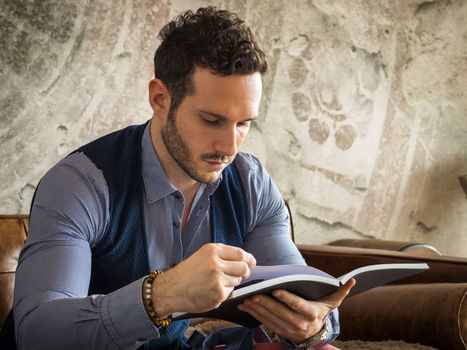 Handsome young man reading book at home in his living-room, sitting on sofa