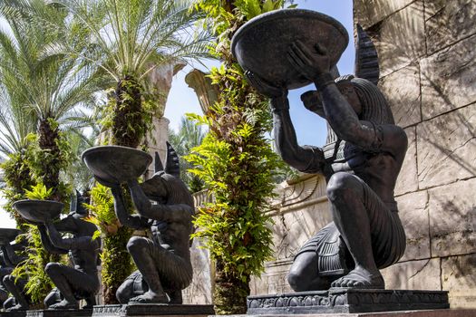 Alley with large identical stone statues of the Egyptian God Anubis, who stands on one knee and holds a round bowl in his hands. Palms grow between the statues, behind them a wall with different symbols.