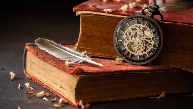 Winding pocket watch on old books with feathers and dried flower petals on the marble table in darkness and morning light.