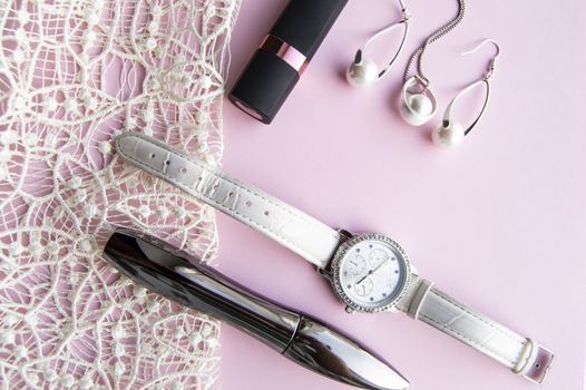 flat lay women's accessories collage with stylish watches, earrings and pendant with white pearls, lipstick, mascara on a Lacy pink background.