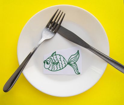 The celebration of April fool's Day, a Plate with a fork and knife and a paper fish on a yellow colored background. Humor.