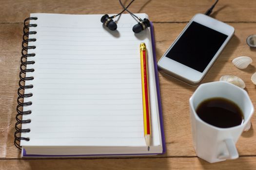 Bank notebook with pencil laying on the brown table. White mobile with earphones and black coffee put on the table as well.