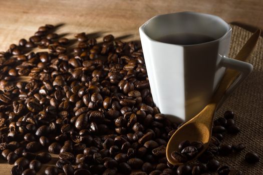 Low key coffee in the darkness. Roasted coffee beans on a wooden table with a glass of white ceramic and a wooden spoon in morning light.