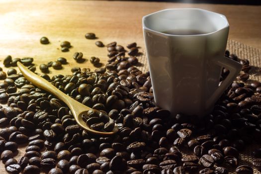 Low key coffee in the darkness. Roasted coffee beans on a wooden table with a glass of white ceramic and a wooden spoon in morning light.