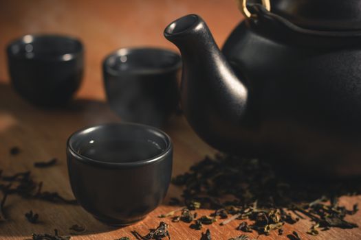 Glass of tea with hot smoke is placed beside a ceramic teapot and dried tea leaves scattered on the wood background.