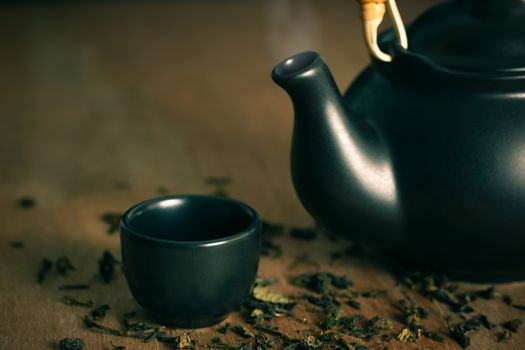 Glass of tea with hot smoke is placed beside a ceramic teapot and dried tea leaves scattered on the wood background.