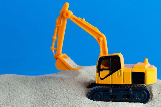 Yellow toy backhoe is digging sand on the blue background. Concept of construction.