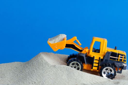 Yellow toy tractor is digging and lift sand on the blue background. Concept of construction.
