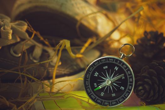 Compass and paper maps with sneakers and pine flowers placed on dry wheat straw in morning sunlight. Concept of adventure tourism or survival in the forest.