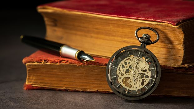 Winding pocket watch placed beside the old book and the pen on the book in darkness and morning light.
