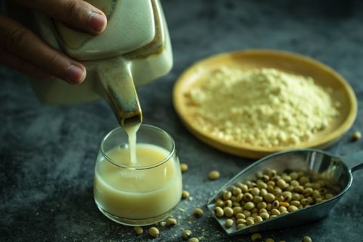 Hand holding the kettle to pour soy milk into the glass. Soybean powder is crushed with soybean seeds scattered on the table and morning light.