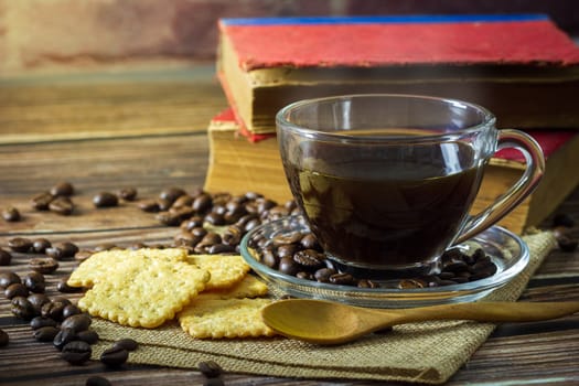 Black coffee in clear glass cup and coffee beans with crackers and old book on wooden table.