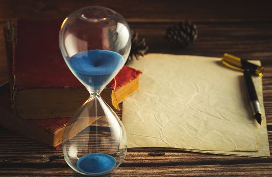 Hourglass and old books with old paper and pen on wooden tables in lighting of the lantern.