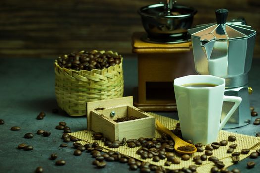 Brew black coffee in white cup and morning lighting. Roasted coffee beans in bamboo basket and wooden spoon. Vintage coffee grinder and pot. Concept of coffee time in morning or start the new day.