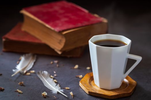 Black coffee in white cup and old books with feather and dried flower petals placed on the marble table and morning sunlight.