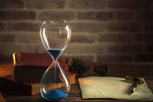 Hourglass and old books with old paper and pen on wooden tables in lighting of the lantern.