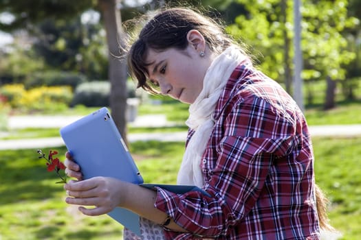 Casual teenage girl in plaid shirt and scarf using tablet and taking picture of small spring flower in sunlight