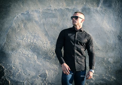 One handsome young man in urban setting in European city, standing, wearing black jacket and jeans