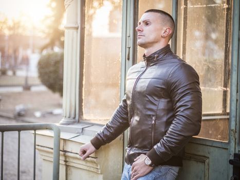 One handsome young man in urban setting in European city, standing, wearing black leather jacket and jeans at sunset