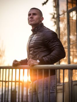 One handsome young man in urban setting in European city, standing, wearing black leather jacket and jeans at sunset