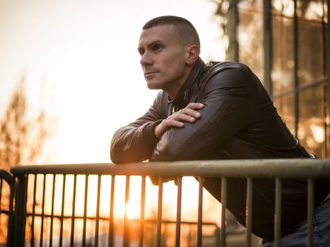 One handsome young man in urban setting in European city, standing, wearing black leather jacket and jeans at sunset