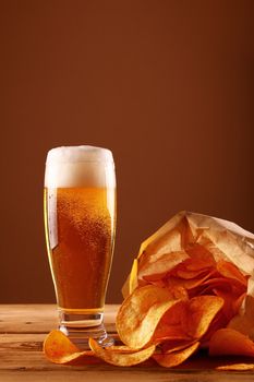 Close up one glass of lager beer with white froth and bubbles and paper bag of potato chips on wooden table over dark brown background with copy space, low angle side view