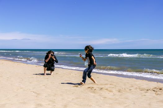 Professional photographer with a camera shoots a running teenager on a sandy beach of the ocean coast on a sunny spring day