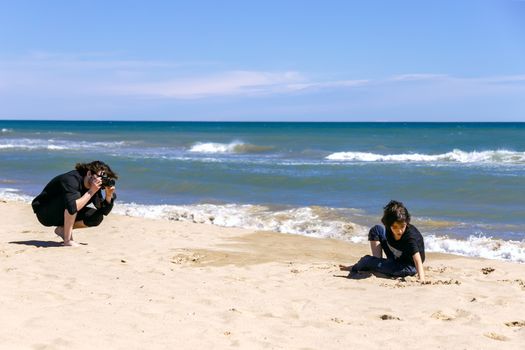 Professional photographer with photo camera shooting teenage boy lying on sandy beach of ocean coast