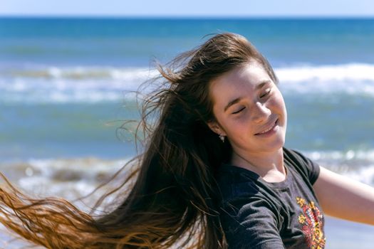 Beauty Sunshine Girl Portrait. A teenager girl with long brown hair flying in the wind smiles happily, closing her eyes and exposing the sun to her face against the backdrop of the sea.