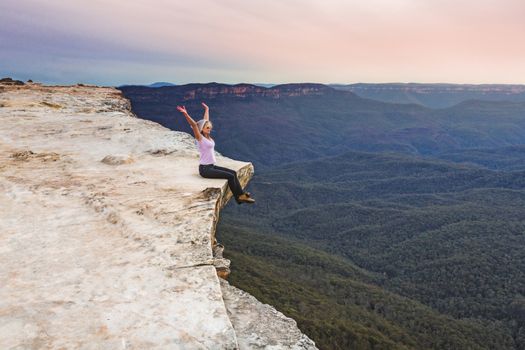 Woman sits on edge of cliff with arms up full of happiness and vitality