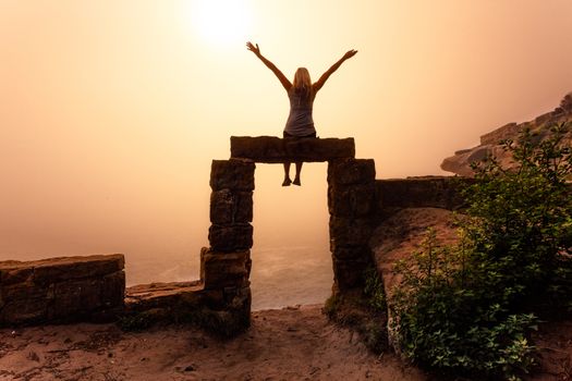 Doorway to the abyss? unknown? future?? Foggy skys and sunlight created a mystical eerie scene. Female sitting on top of old hand hewn sandstone door arch on the sea coast cliff edge facing into the unknown