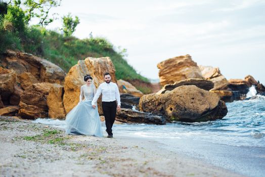 same couple with a bride in a blue dress walk along the ocean shore