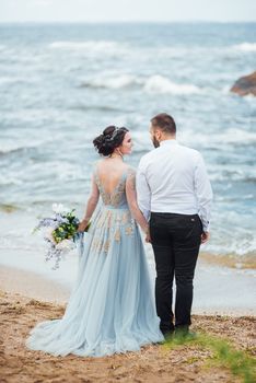 same couple with a bride in a blue dress walk along the ocean shore