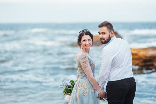 same couple with a bride in a blue dress walk along the ocean shore